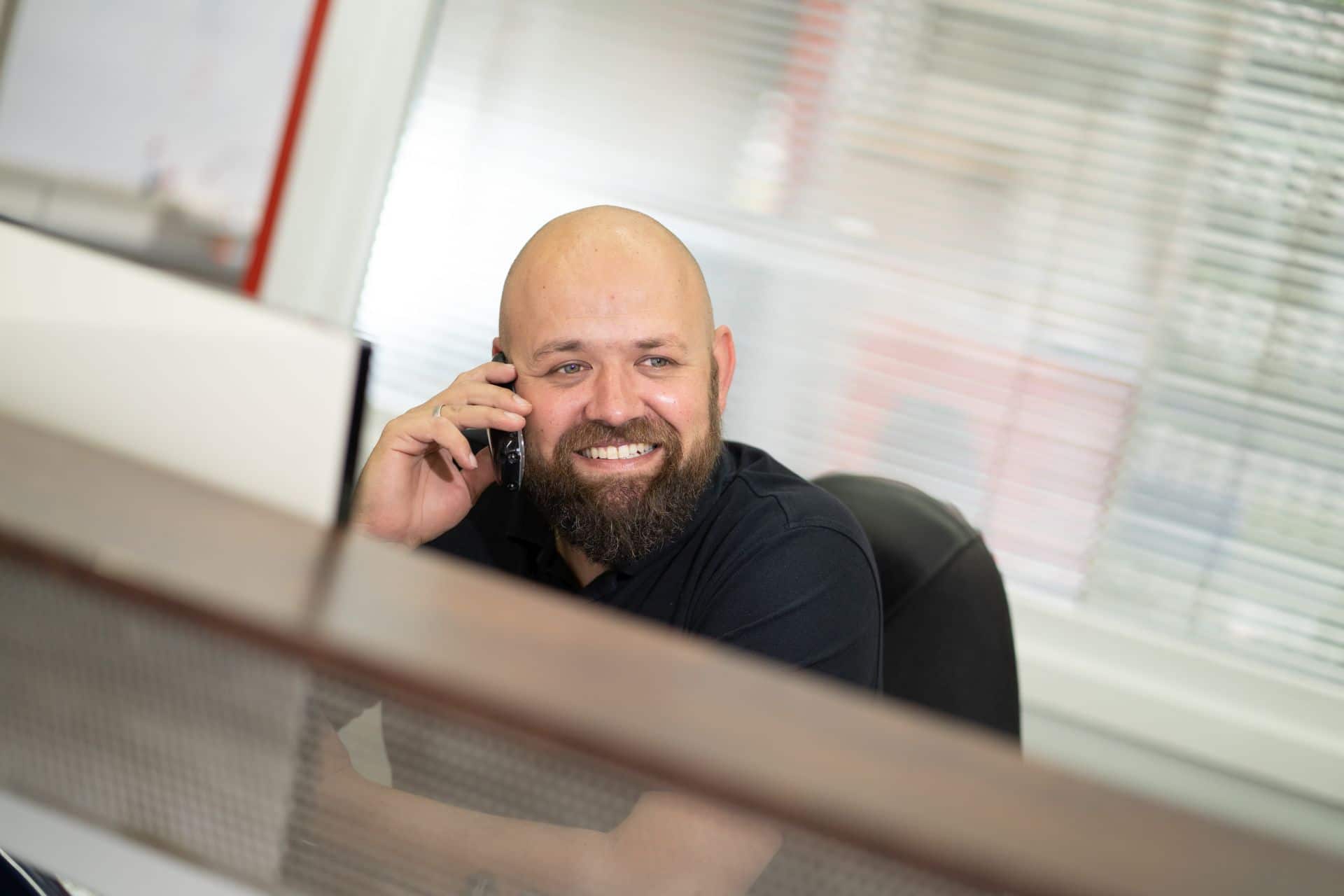 Ein lächelnder, bärtiger Mann mit Glatze sitzt am Schreibtisch und telefoniert in einem modernen Büro mit Jalousien im Hintergrund.