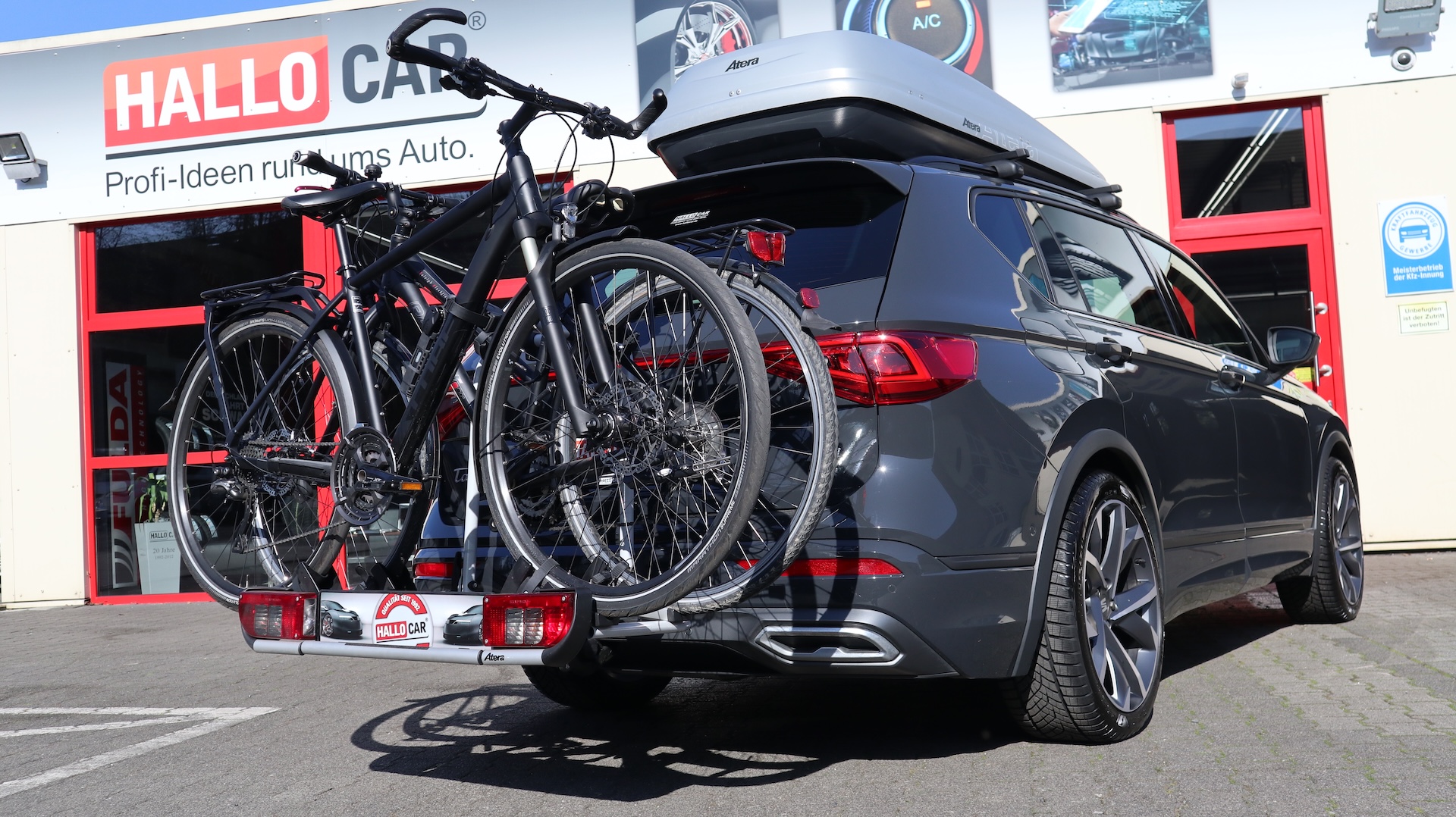 Grauer Kombi mit zwei montierten Fahrrädern am Heckträger und einem großen weißen Dachkoffer, geparkt vor einem Gebäude mit rotem Türrahmen und einem Schild "HALLO CAR".