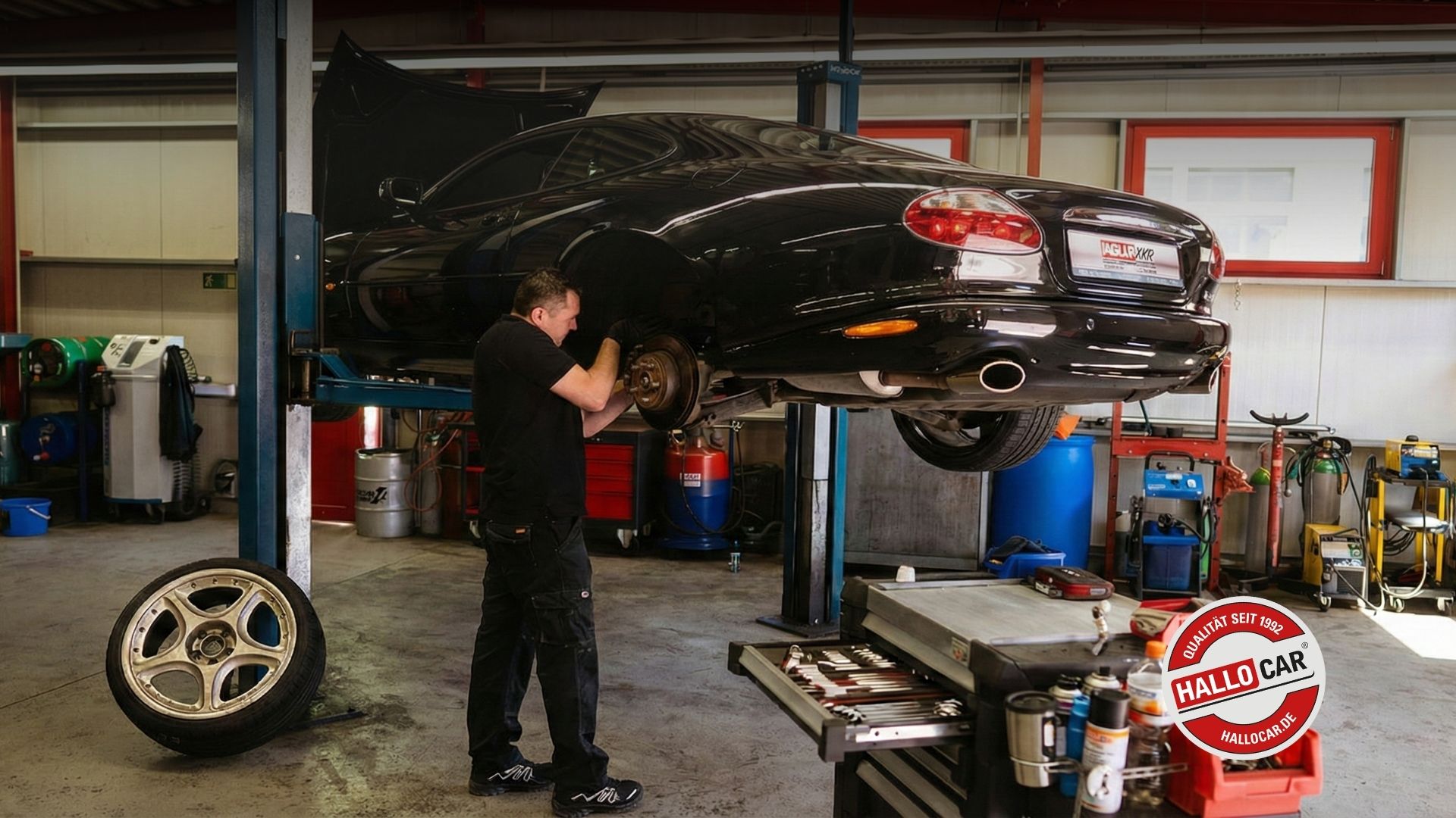 Mechaniker repariert schwarzen Jaguar XKR auf Hebebühne in Werkstatt, Rad abgenommen und Werkzeugwagen im Vordergrund sichtbar.